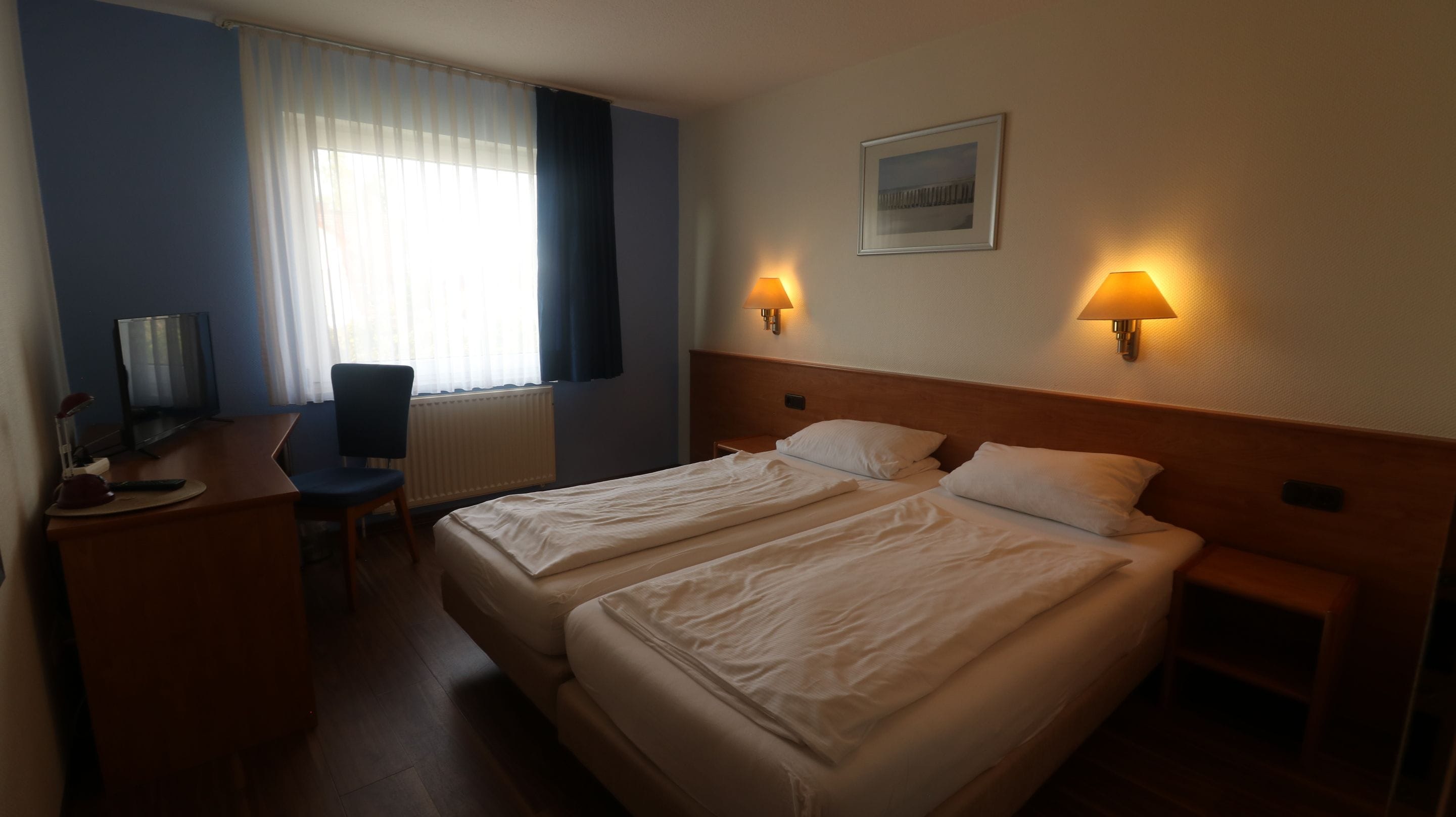 Habitación de hotel con dos camas, un escritorio, una silla, un televisor y dos lámparas de pared.