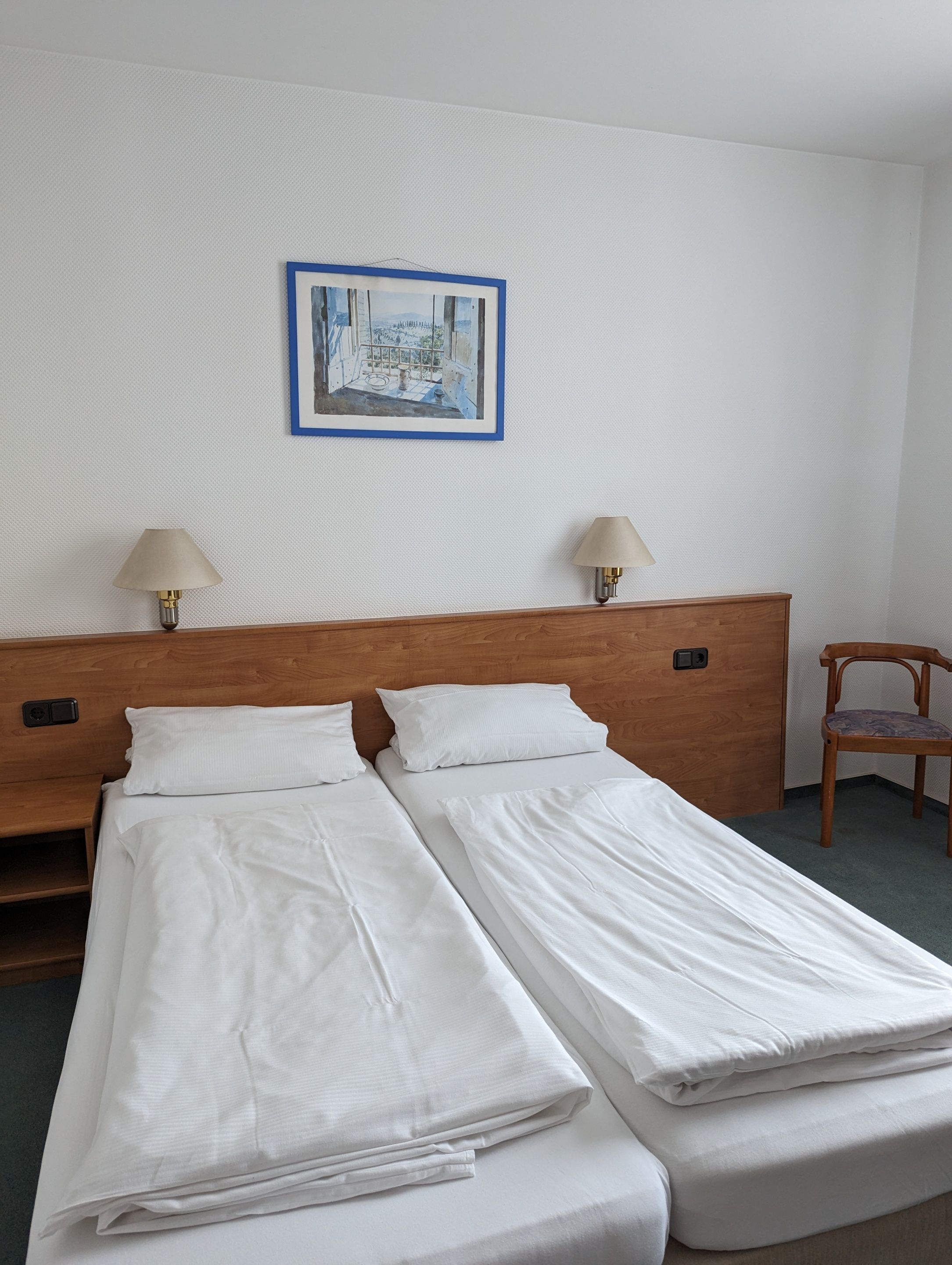 Una habitación de hotel con una cama doble, dos lámparas de noche, un cabecero de madera, un cuadro enmarcado en la pared y una silla de madera.