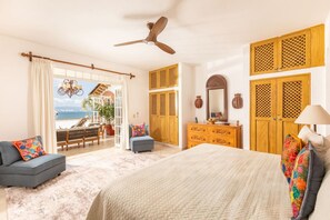 Ein Schlafzimmer mit Bett, Schminktisch, Stuhl, Deckenventilator und Blick auf das Meer.