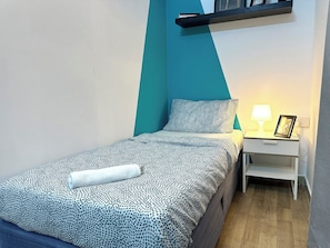 Een klein, netjes opgemaakt bed met een bedovertrek met patroon en kussens, een nachtkastje met een lamp en een fotolijstje, en een plank boven het bed.
