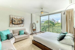 A modern bedroom with a large bed, a sofa, a ceiling fan, and a view of greenery outside. - 3BR Playa Condo: Rooftop Pool, Steps to 5th Ave (Playa del Carmen)