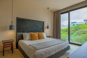 A modern hotel room with a large bed, two bedside tables, a large window with a view of greenery, and a dark headboard. - Beachfront 2BR Rooftop Pool Playa (Playa del Carmen)