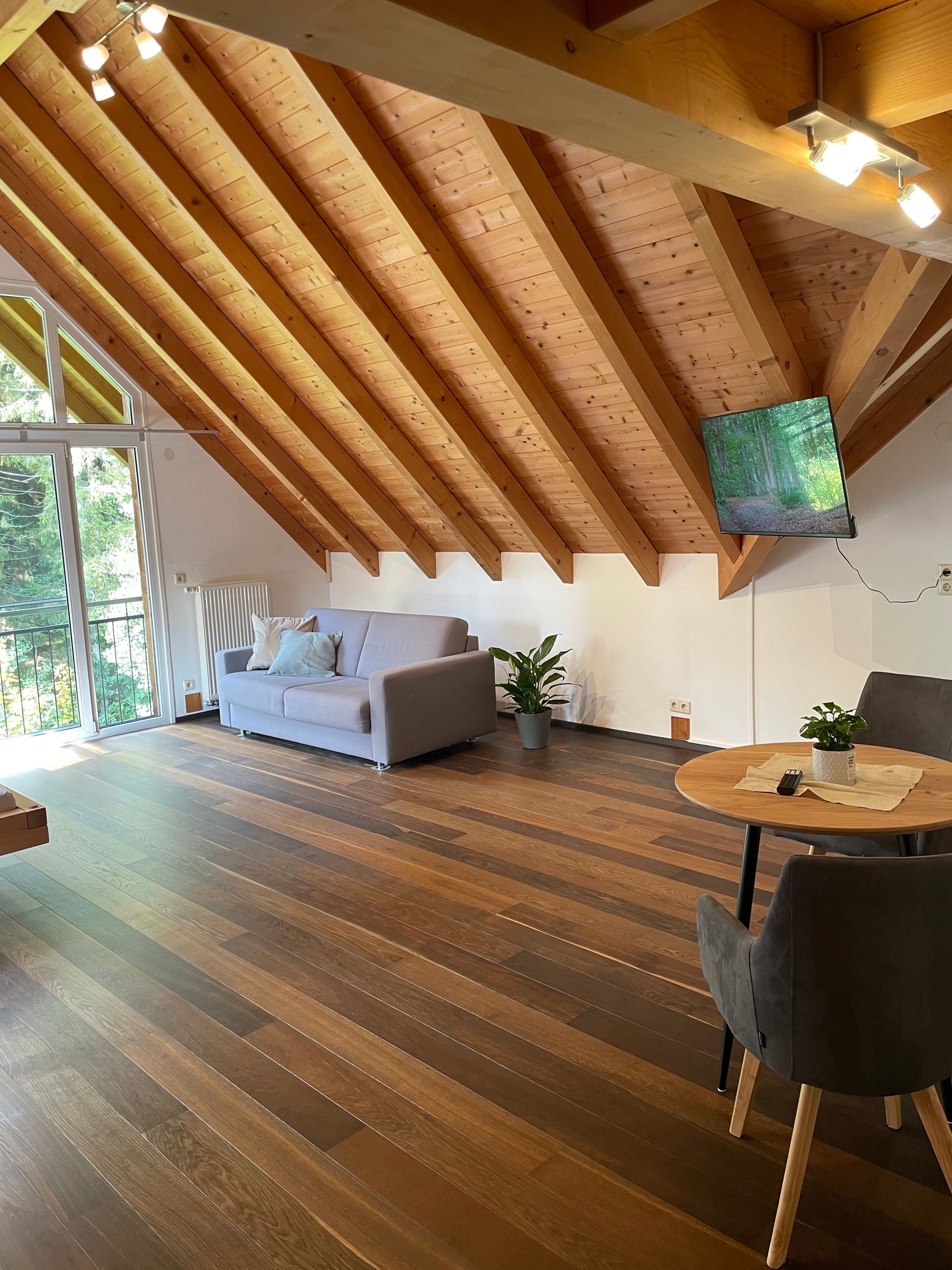 A modern living room with a sloped ceiling, wooden beams, a grey sofa, a round wooden table, and a television mounted on the wall.