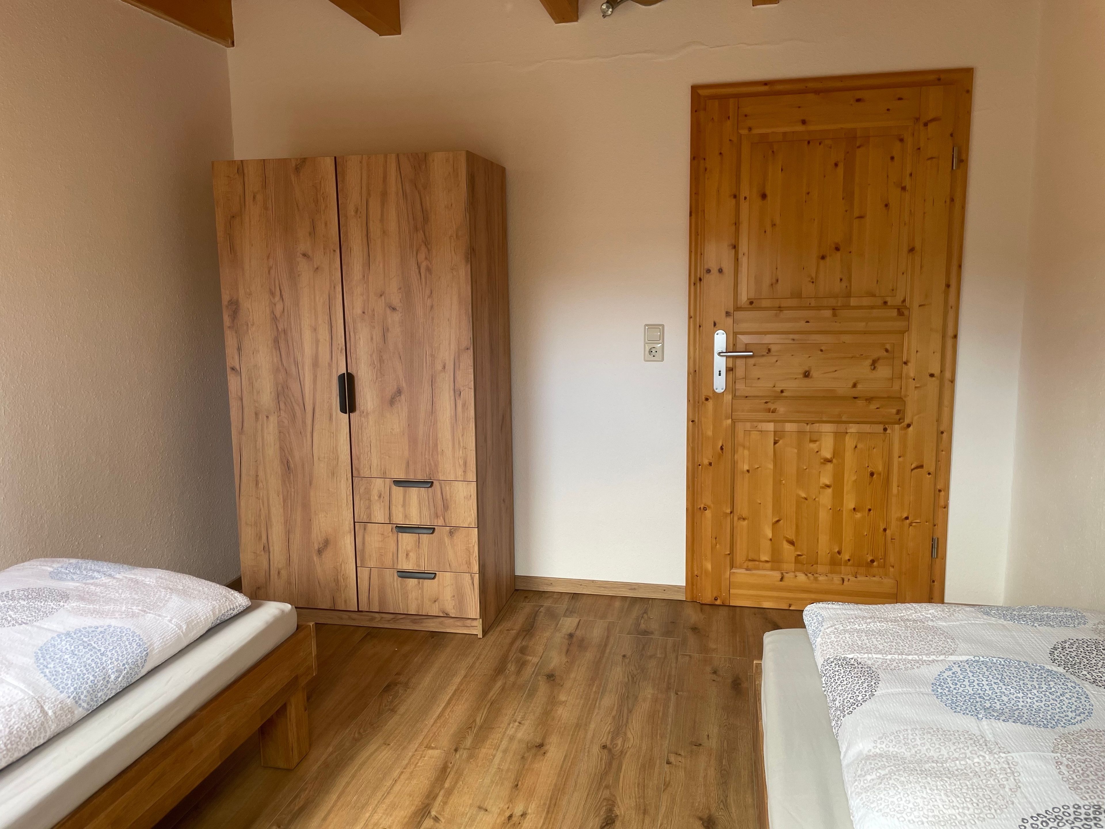 A room with a wooden wardrobe, two beds with patterned bedding, and a wooden door.