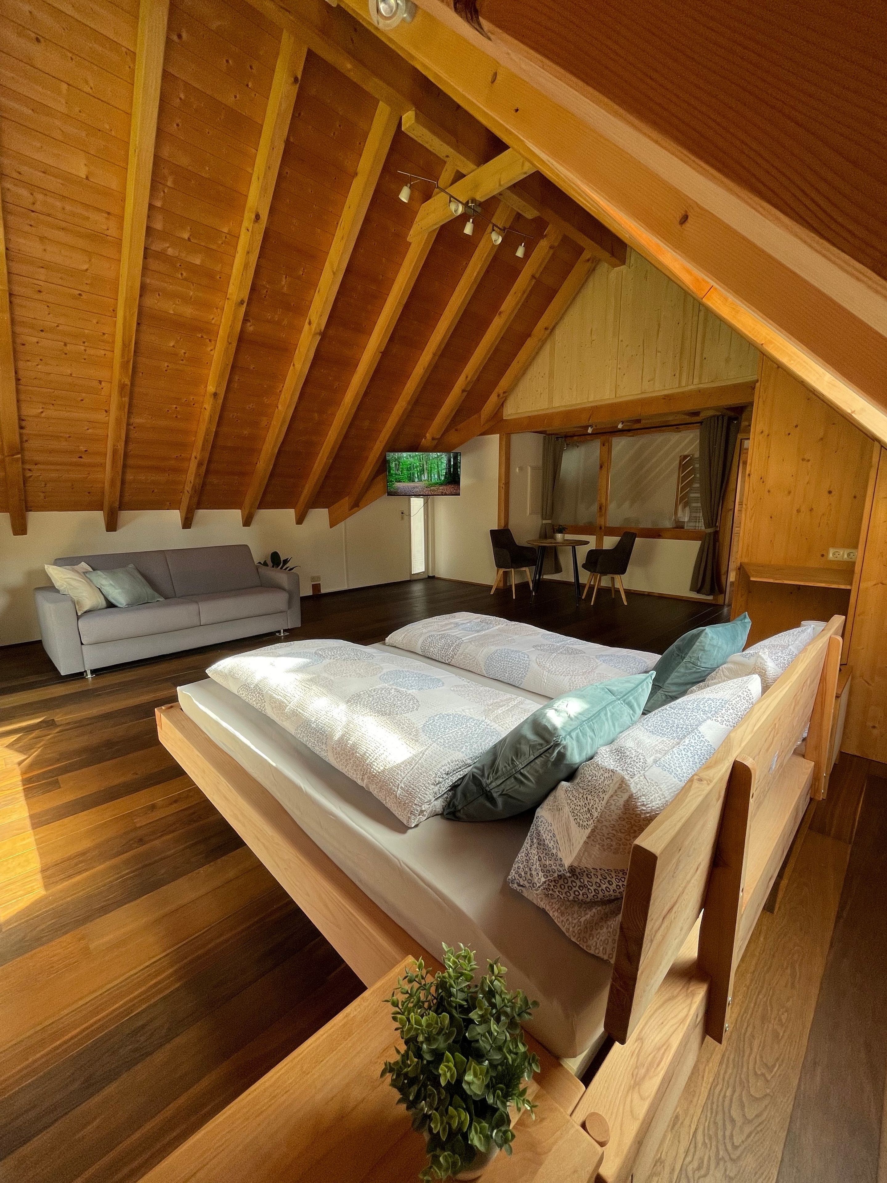 Ein gemütliches Schlafzimmer im Dachgeschoss mit Holzbalkendecke, einem Bett mit gemusterten Textilien, einem grauen Sofa und einem Fernseher an der Wand.