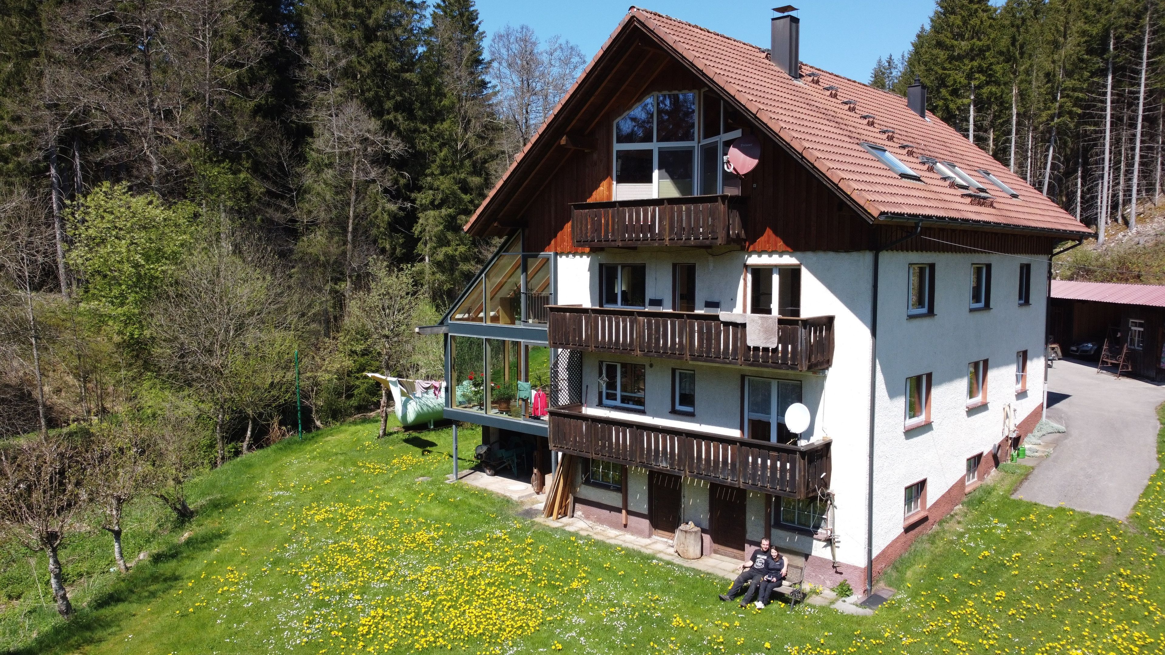 Un chalet à deux étages doté d’un balcon, entouré d’une zone boisée et d’une pelouse parsemée de fleurs jaunes.