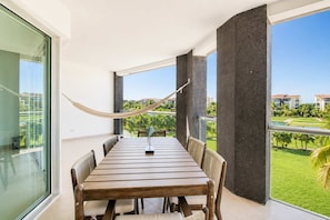 A balcony with a wooden table and chairs, a hammock, and a view of a green landscape. - Beachfront 2BR Condo, Resort Pool, Gym & Beach (Playa del Carmen)