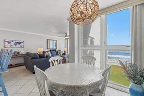 Ein Essbereich mit einem Tisch, der mit einer strukturierten weißen Tischdecke bedeckt ist, einem Deckenventilator und Blick auf das Meer durch große Fenster.
