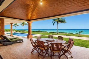 A covered outdoor area with a pool, dining table, and chairs, overlooking a beach with palm trees and the ocean. - Beachfront 5BR Villa Playacar, Private Pool (Playa del Carmen)