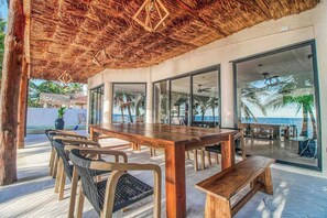 An outdoor dining area with a wooden table and chairs, a thatched roof, and large windows offering a view of the ocean and palm trees. - Beachfront Home Private Pool, Jacuzzi, Sauna (Tulum)