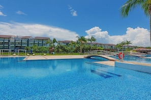 A swimming pool with a bridge, palm trees, and hotel buildings in the background. - Beachfront Studio Resort Pool & Beach Access (Playa del Carmen)