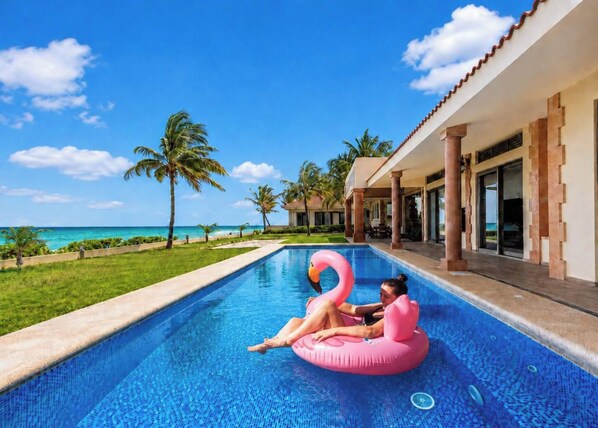 A swimming pool with a clear view of the ocean and palm trees, a person on a pink float, and a building with a tiled roof. - Beachfront Villa Private Chef, Pool & Transfer (Playa del Carmen)