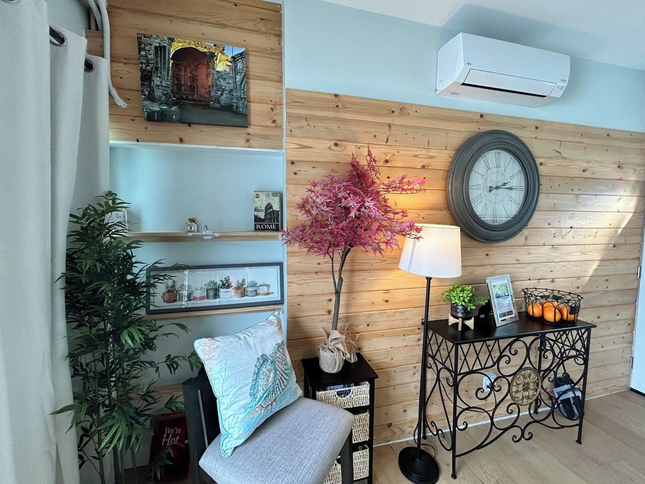 Una habitación con pared de madera, un reloj redondo, una lámpara, una pequeña mesa con adornos y una silla con cojín.