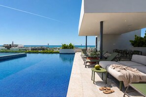 A modern outdoor pool area with lounge chairs and a sofa, overlooking a clear blue sky and a distant view of buildings. - Ceiba 25 Rooftop Pool, Gym & 5th Ave Condo (Playa del Carmen)