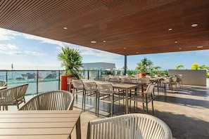 An outdoor seating area with tables and chairs, overlooking a body of water and a distant building. - Ceiba 25 Rooftop Pool, Gym & 5th Ave Condo (Playa del Carmen)