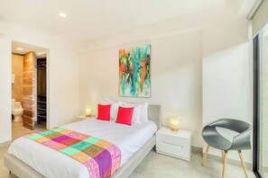 A modern bedroom with a large bed, a colorful quilt, a nightstand with a lamp, a chair, and a bathroom visible through an open door. - Icono Ocean-view Condo Rooftop Pool, Playa (Playa del Carmen)