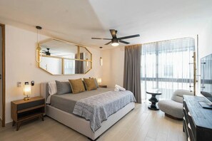 A modern hotel room with a large bed, a ceiling fan, a bedside table with a lamp, a chair, and a view of the city through large windows. - Luxury 3BR Penthouse w/ Pool Near Beach & 5th Ave (Playa del Carmen)