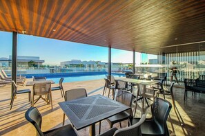 An outdoor terrace with tables and chairs, overlooking a swimming pool and cityscape. - Luxury Condo: Rooftop Pool, Jacuzzi & Game Room (Playa del Carmen)