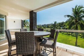 A balcony with a round table and wicker chairs, overlooking a well-maintained lawn and palm trees. - Mareazul 516n Beachfront 3BR Condo Playa (Playa del Carmen)