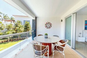 A balcony with a round table and chairs, a decorative wall piece, and a view of a garden and buildings. - Mareazul Beachfront 2BR Pool & Sea Views (Playa del Carmen)