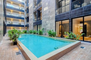 A modern indoor swimming pool with a tiled floor, surrounded by potted plants and a multi-level building with balconies. - Modern 1BR Condo Rooftop Pool & Gym, Near Beach (Playa del Carmen)
