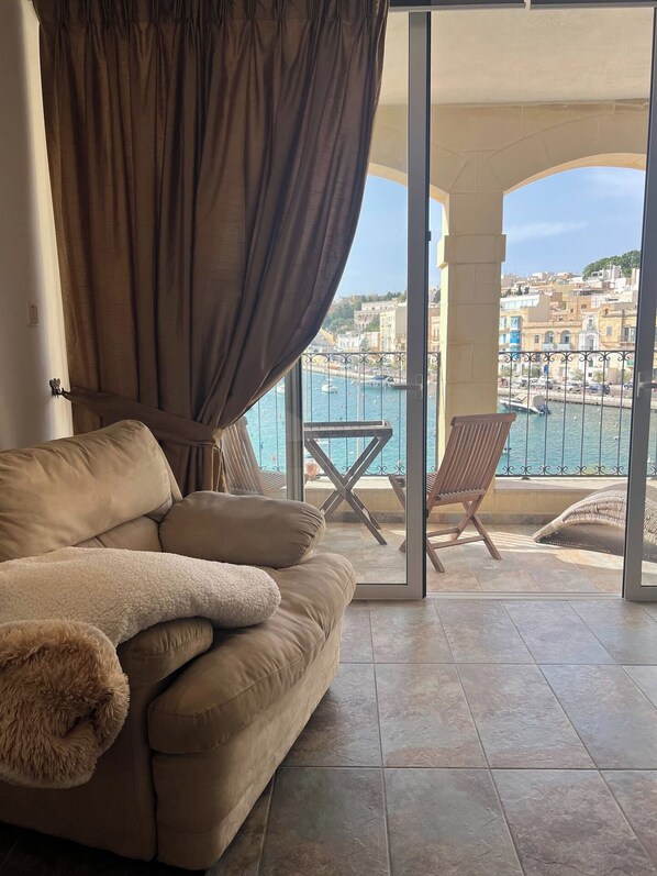 A room with a beige sofa, a fluffy pillow, and a view of a coastal city through a glass door.