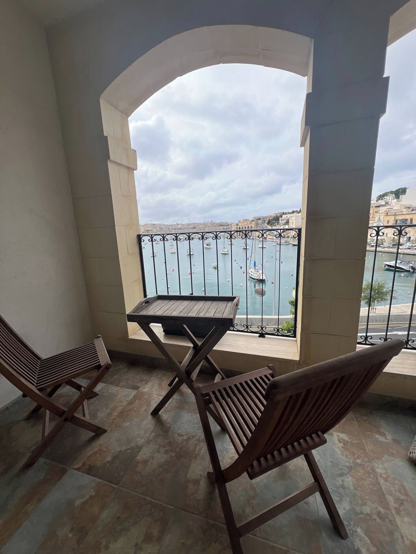 A balcony with a table and two chairs, overlooking a body of water with boats and buildings in the background.