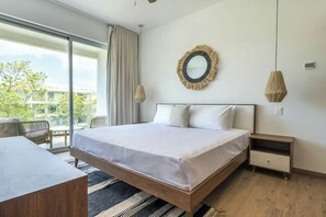 A hotel room with a bed, a nightstand, a bedside table, a large window, and a view of greenery outside. - Modern 2BR Condo: Pool View, Kitchen & Wifi (Playa del Carmen)