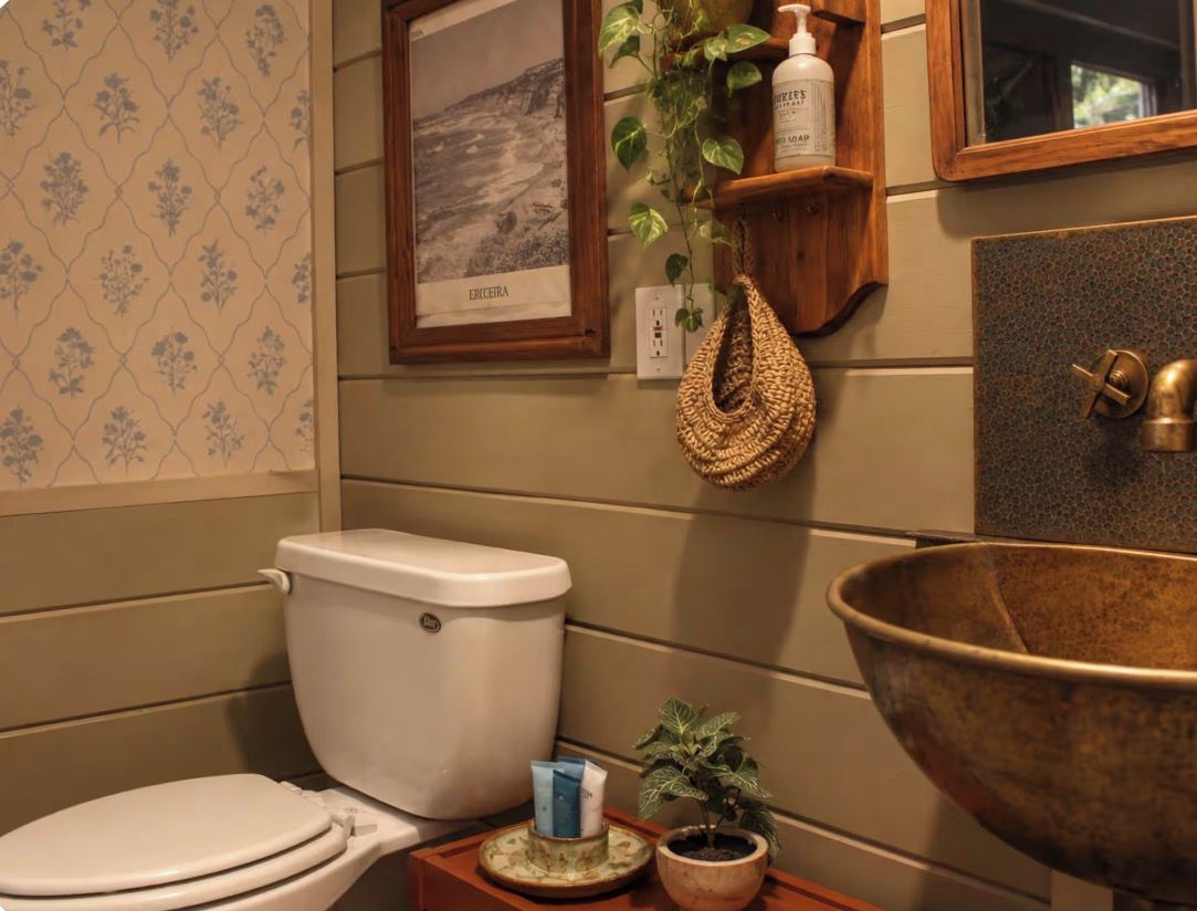 A bathroom with a toilet, a sink with a large basin, a framed picture on the wall, and a shelf with toiletries.