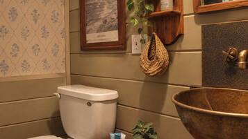 A bathroom with a toilet, a sink with a large basin, a framed picture on the wall, and a shelf with toiletries.