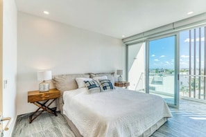 A bedroom with a large bed, a nightstand, a lamp, and a view of the city through a large window. - Ocean View Condo w/ Luxury Pool Rooftop Access (Cancun)