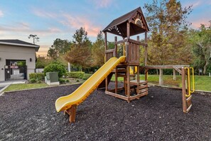 A playground with a slide and climbing structure, surrounded by a landscaped area with trees and a building in the background. - Terra Esmeralda Home Near Disney for 9 Guests (Kissimmee)