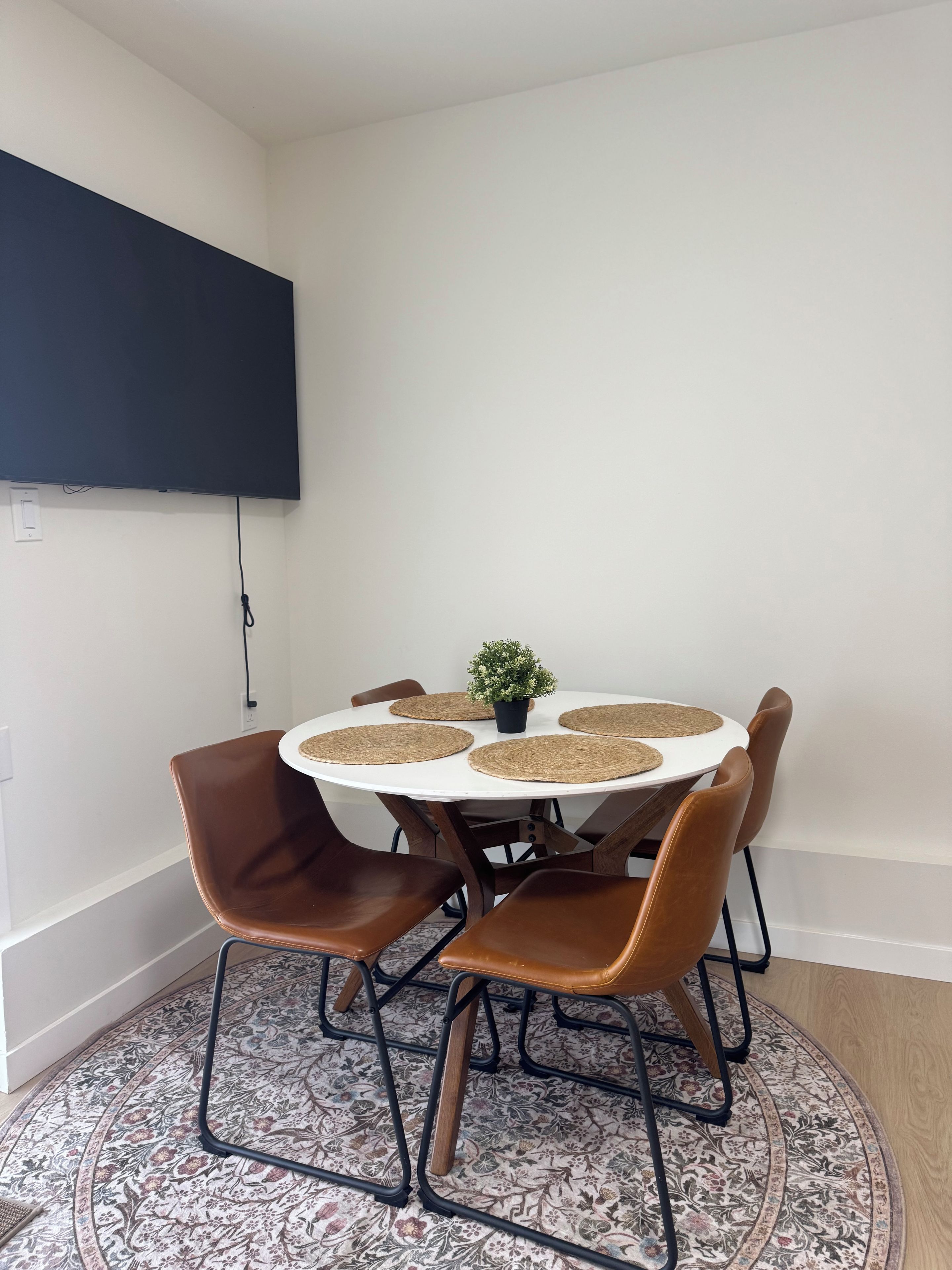 Un comedor con una mesa redonda, cuatro sillas, una pequeña planta en maceta y un televisor de pantalla plana montado en la pared.