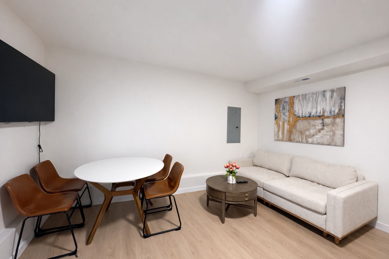 A modern living room with a round dining table, two brown chairs, a beige sofa, a small round coffee table with a vase of flowers, and a mounted artwork on the wall.