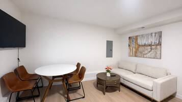 A modern living room with a round dining table, two brown chairs, a beige sofa, a small round coffee table with a vase of flowers, and a mounted artwork on the wall.