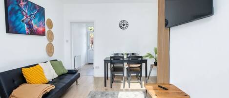 A modern living room with a black sofa, a dining table, and a TV mounted on the wall.
