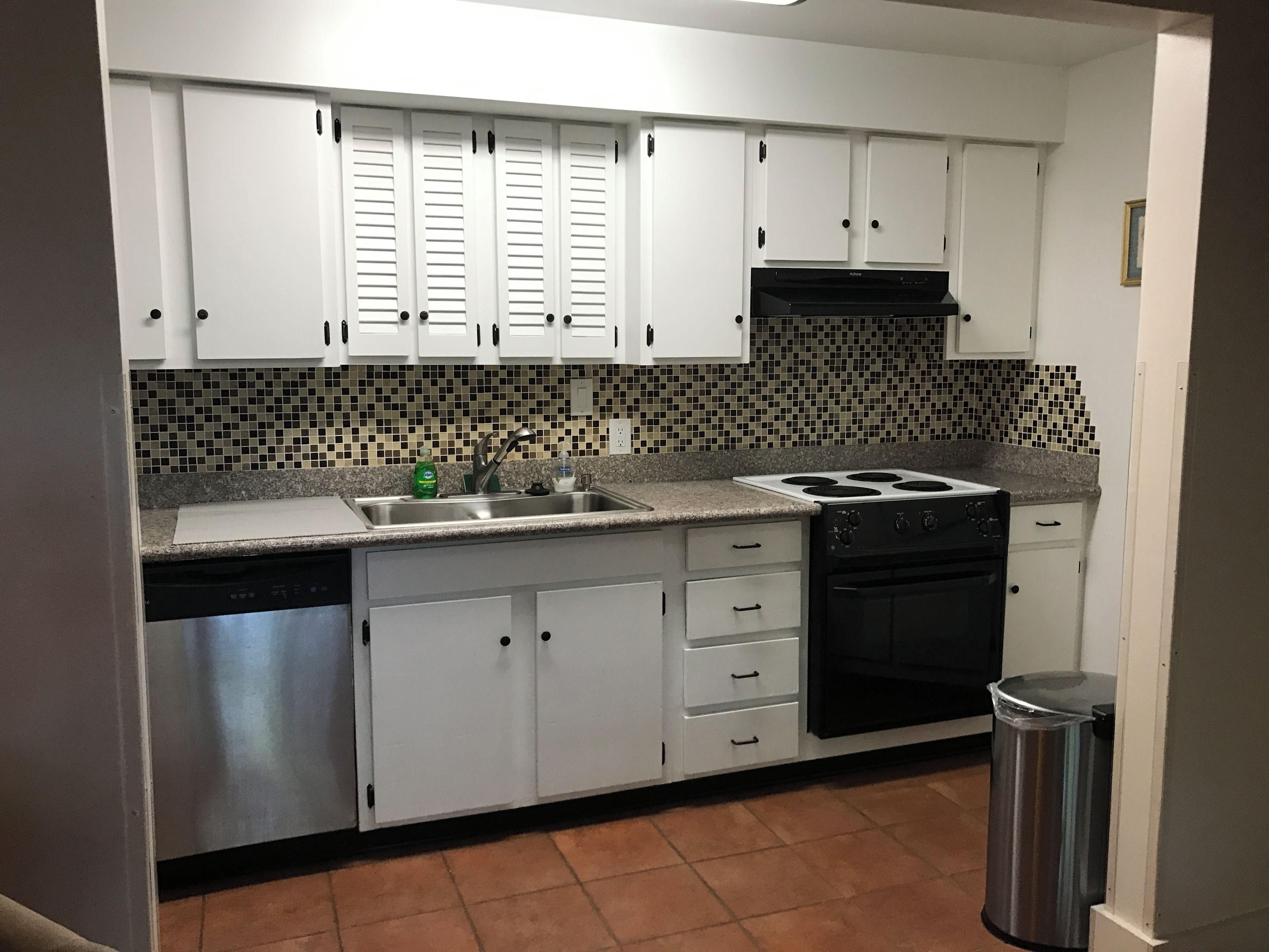A kitchen with white cabinets, a tiled backsplash, a stainless steel sink, a stove, and a trash can.