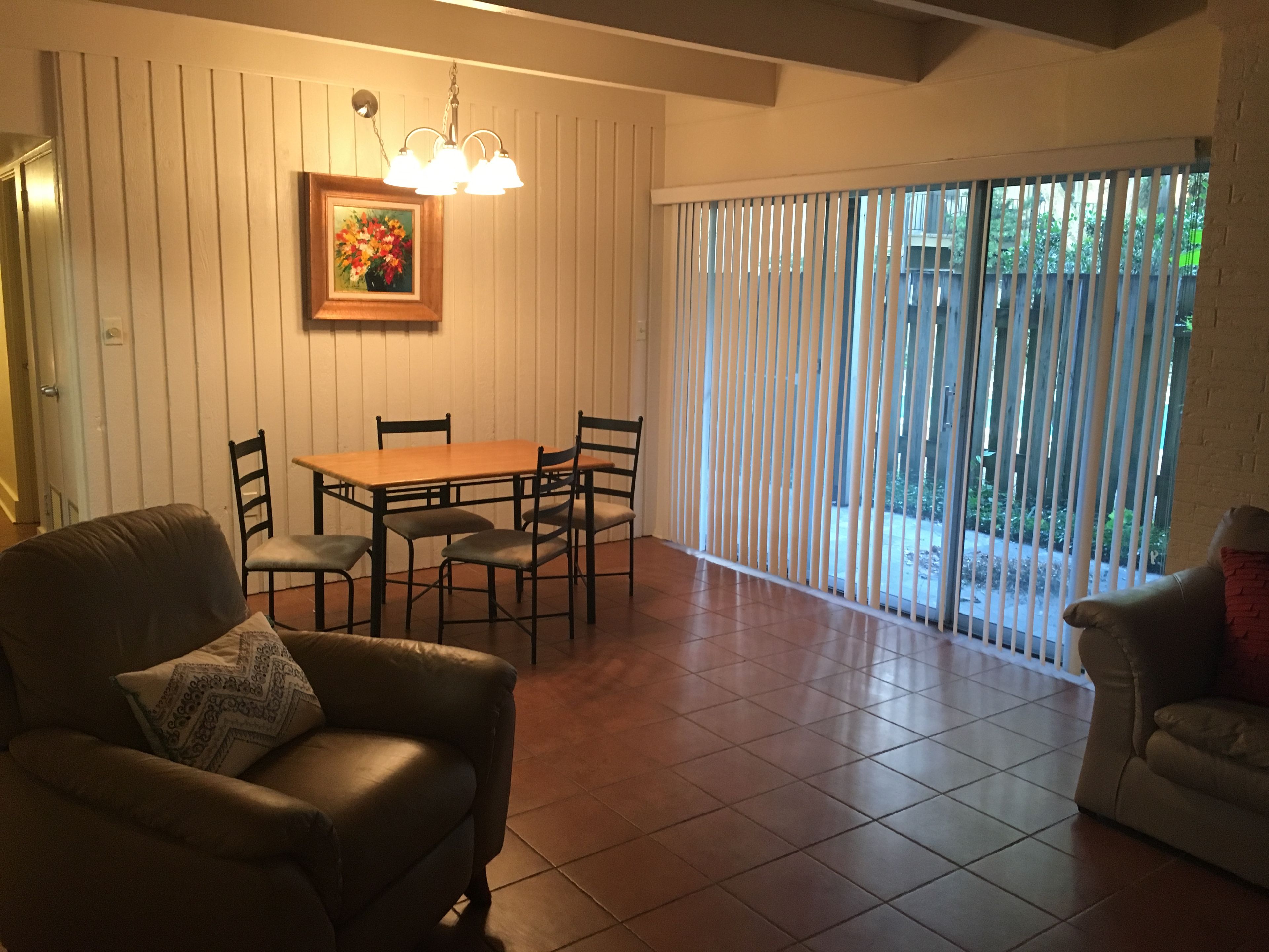 Un comedor con mesa y sillas de madera, un cuadro en la pared y un cerramiento de vidrio corredero que da a un espacio exterior.