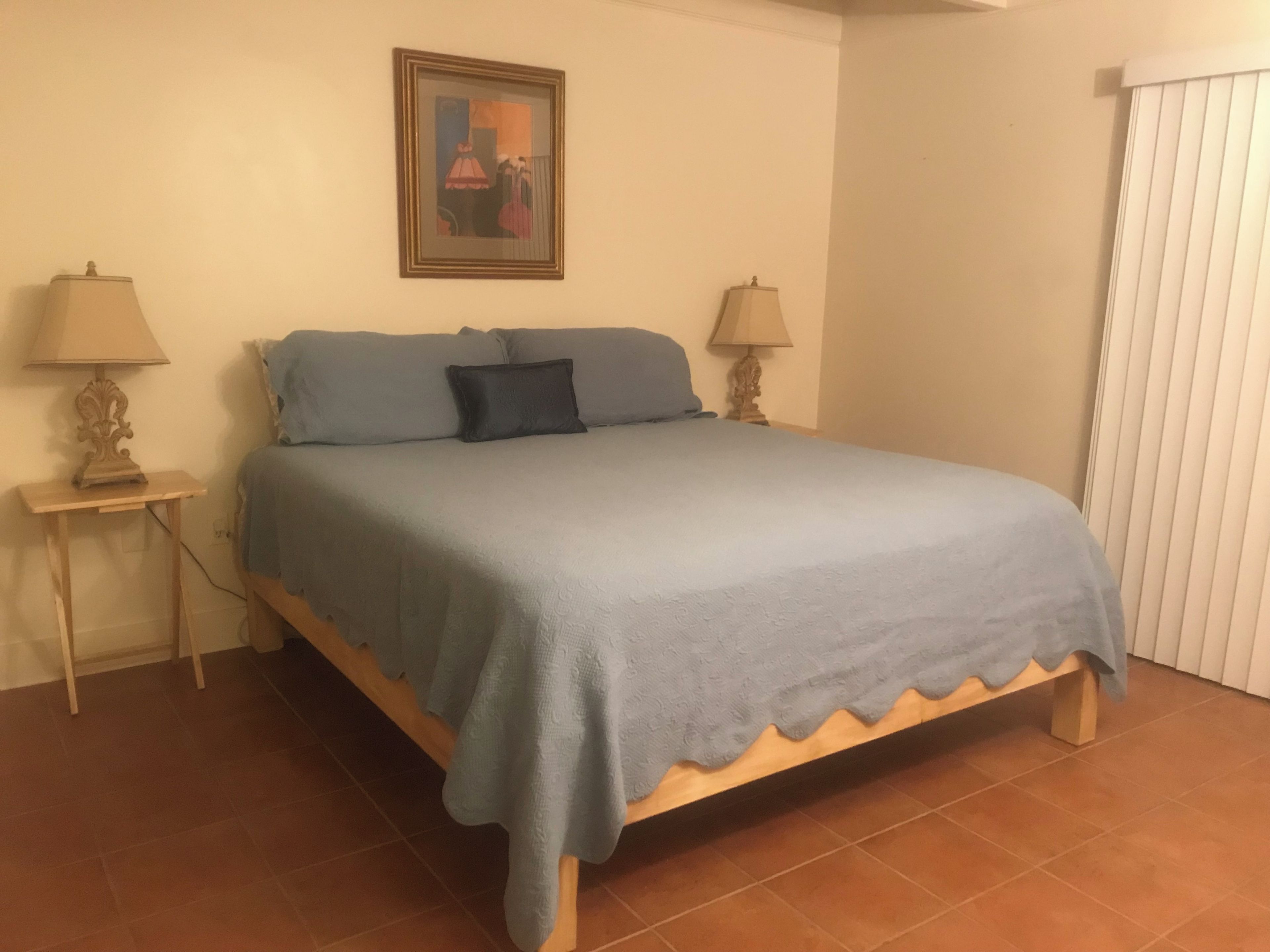 Un dormitorio con cama, dos mesitas de noche, una lámpara sobre cada mesita y un cuadro en la pared, además de una ventana con persianas.