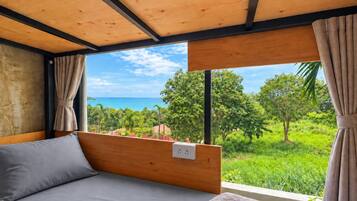 A bedroom with a bed, a window with curtains, and a view of greenery and the sea.