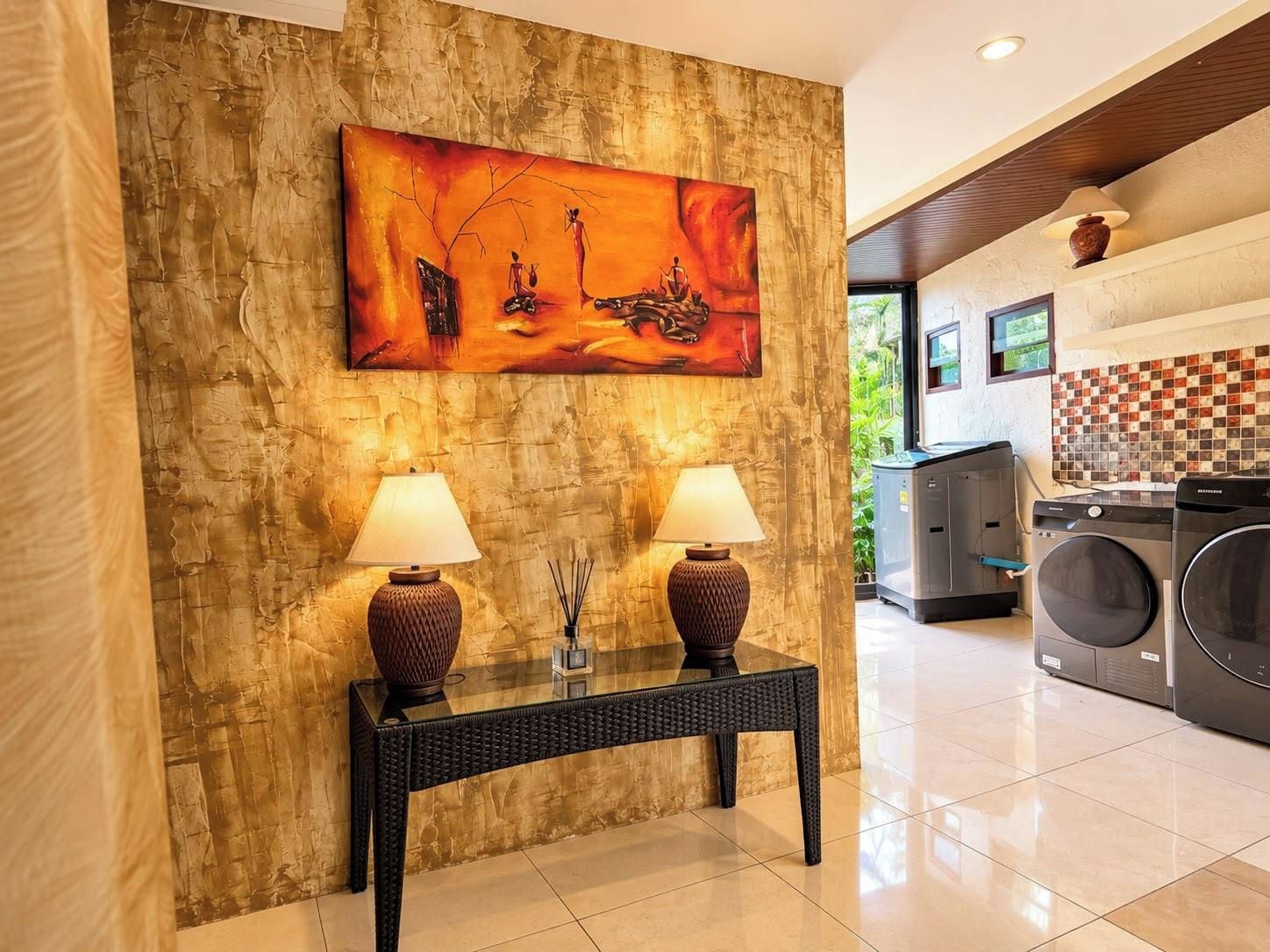 Vestíbulo de hotel con un cuadro abstracto grande, dos lámparas sobre una consola y un área de lavandería con lavadoras.