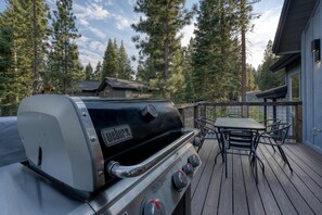 A Weber grill on a wooden deck with a table and chairs, surrounded by tall trees. - Tahoe Vista Sanctuary | Hot Tub + Fireplace (Tahoe Vista)