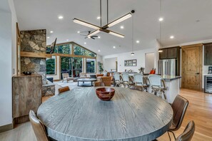 Un vaste espace de vie comprenant un coin repas avec une table ronde, une cheminée en pierre et une grande fenêtre donnant sur une forêt.