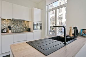 A modern kitchen with white cabinetry, a tiled backsplash, a built-in oven, and a large window offering a view of the city. - SUDANVERS (Antwerpen)