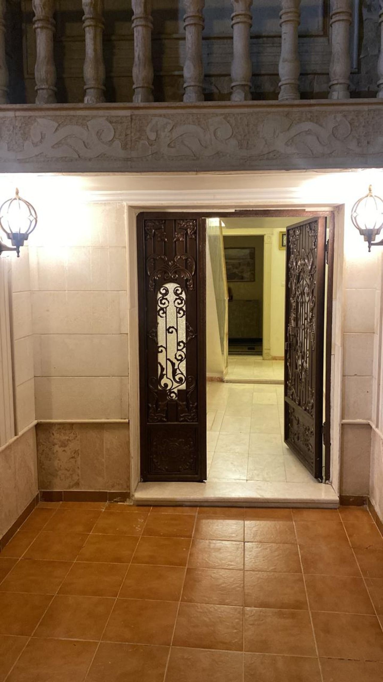 Une porte surmontée d’un portail en métal décoratif, qui donne sur un couloir carrelé avec un escalier.