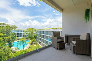 A balcony with wicker furniture, a hanging plant, and a view of a swimming pool and building. - Playacar 2BR Balcony, Pool & Beach Access (Playa del Carmen)