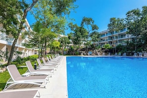 A swimming pool with lounge chairs, surrounded by trees and a building with multiple floors. - Playacar Condo - Balcony - Pools - Sleeps 6 (Playa del Carmen)