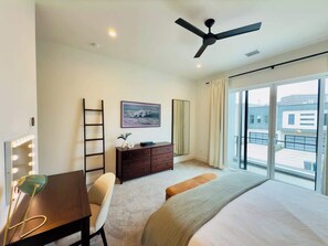 A hotel room with a bed, a desk with a lamp, a dresser, a mirror with lights, a ceiling fan, and a large window with a view of buildings. - Peaceful Dallas Oasis: Fireside Patio, Chef's Kitchen, King Beds, Dogs Welcome (Dallas)