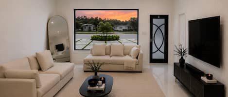 Una sala de estar moderna con un sofá, una mesa de centro, un televisor y un ventanal con vistas a una zona residencial.