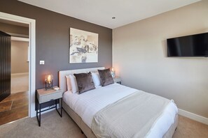 Ein Schlafzimmer mit einem Bett, Nachttischen, einem Gemälde, einem Fernseher und einer Tür zu einem anderen Raum.